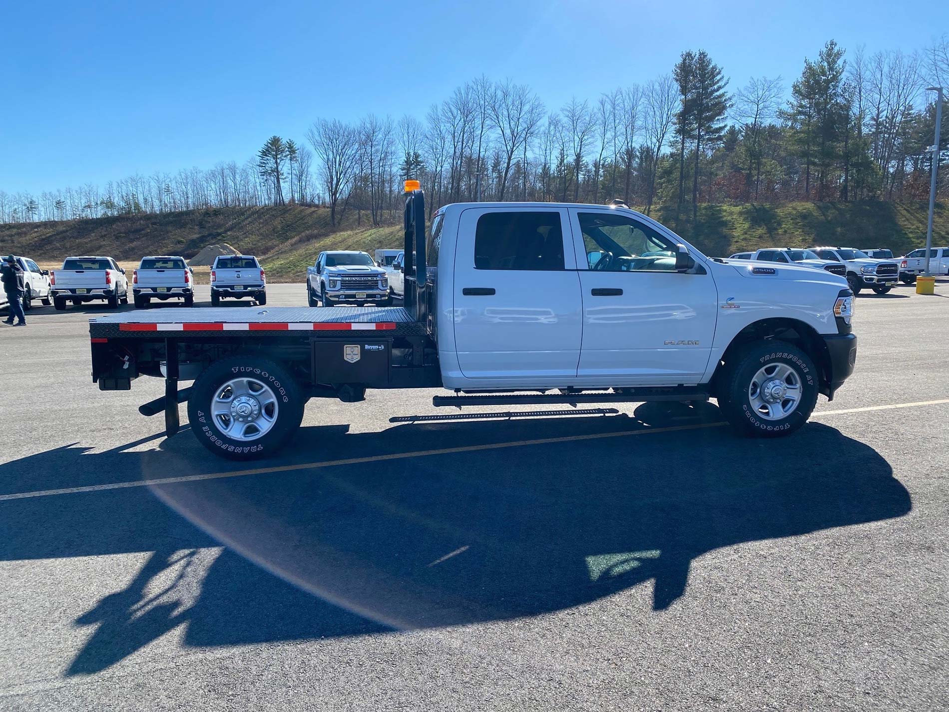 White flatbed truck parked in a sunny lot, surrounded by other vehicles and trees in the background.