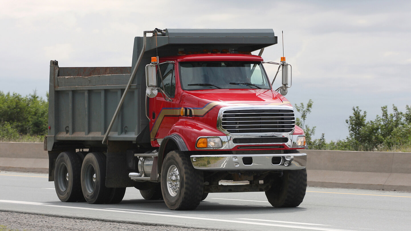 Red dump truck driving on a highway, with trees and a cloudy sky in the background.