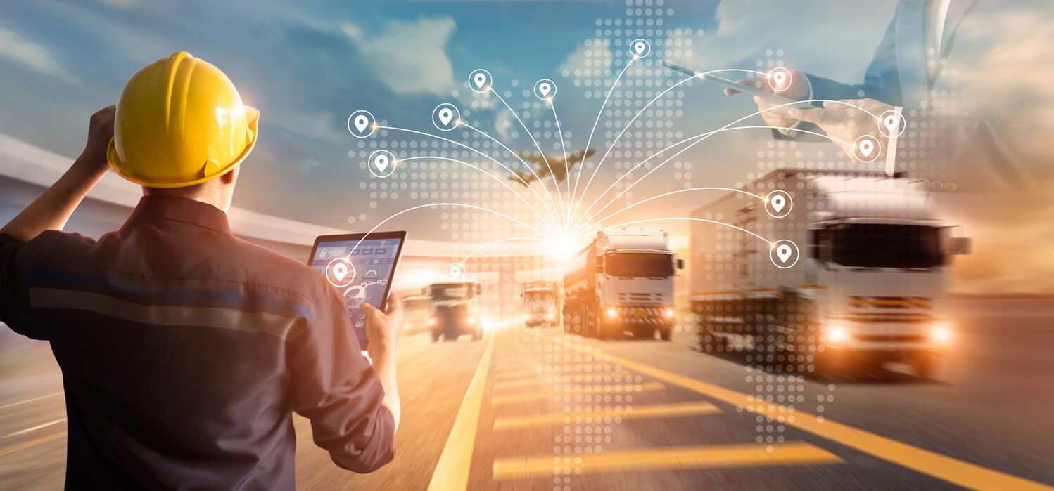A worker with a hard hat and a tablet observes trucks on a road with a digital map overlay showing connections.