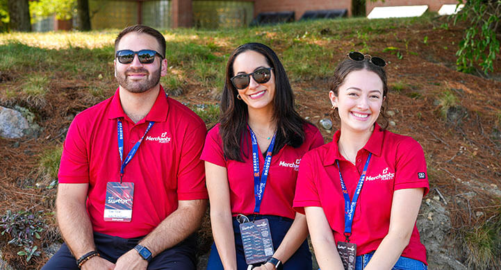 Three people in red shirts with name tags sit outdoors, smiling at the camera.