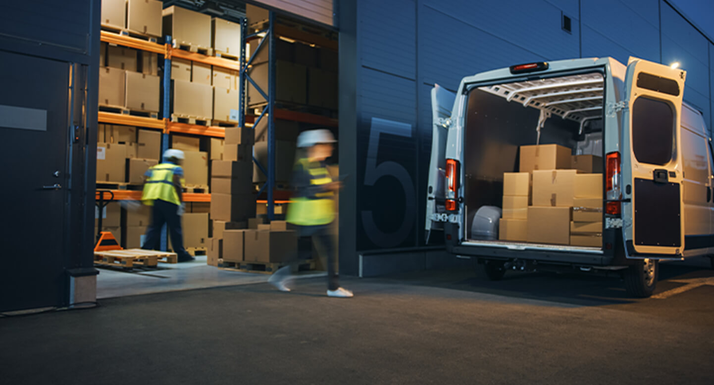 Workers load boxes into a white van at a warehouse loading dock at night.