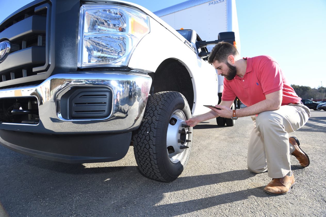 Man in red shirt crouches beside a truck tire, examining it closely in a sunny outdoor setting.